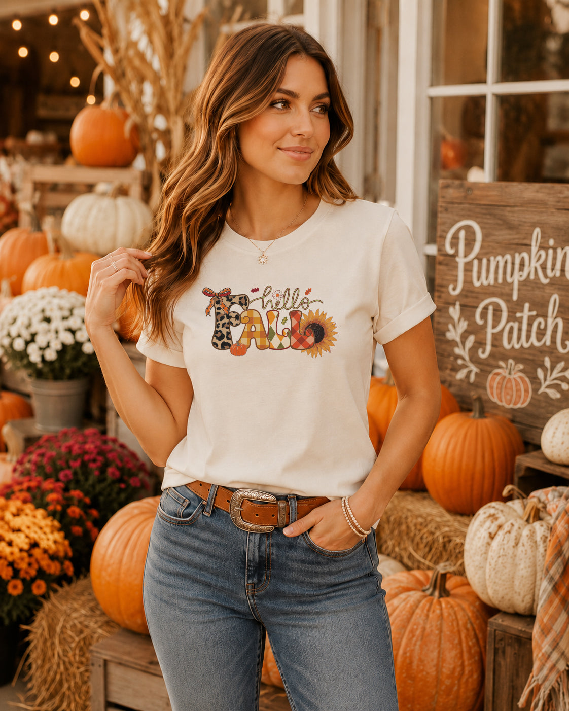 A woman wearing a cream graphic t-shirt with a 'Hello Fall' autumn-themed design, paired with blue jeans, posed in front of a rustic fall display with pumpkins and a 'Pumpkin Patch' sign.