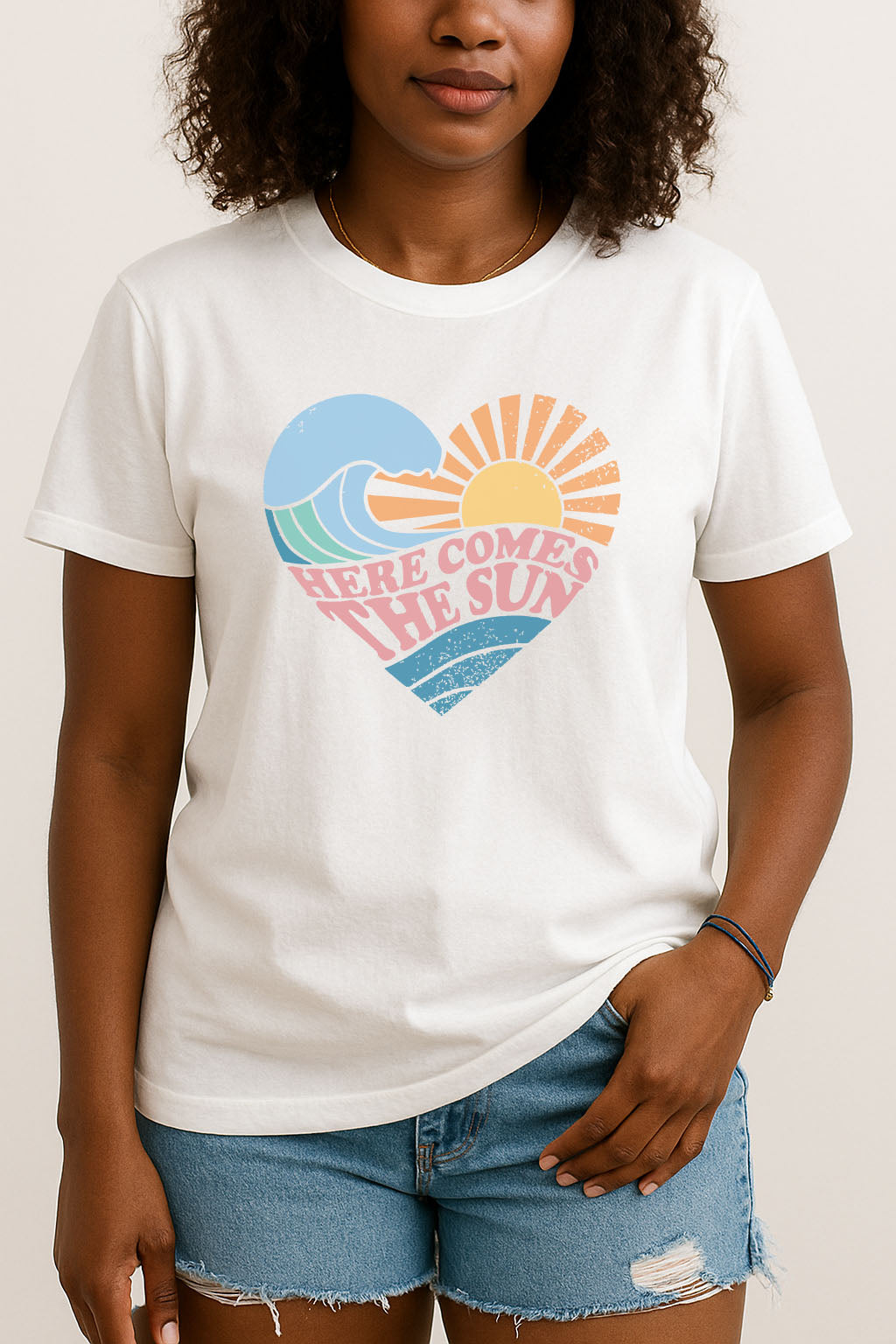 Model wearing white women's retro graphic tee with pastel heart illustration, waves, sunshine, and ‘Here Comes The Sun’ lettering.