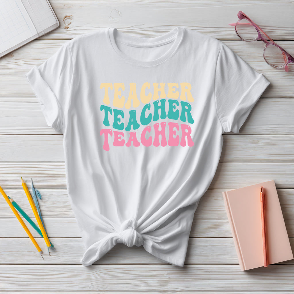 White t-shirt with 'TEACHER TEACHER TEACHER' text in pastel colors on a wooden surface with stationery items.