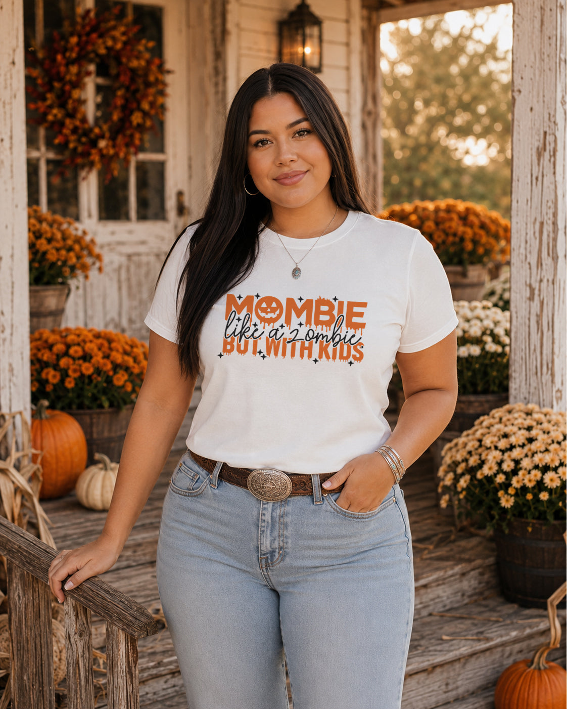 Woman wearing the Mombie Like a Zombie But With Kids Halloween mom graphic tee on a cozy fall porch with pumpkins, mums, and autumn décor.