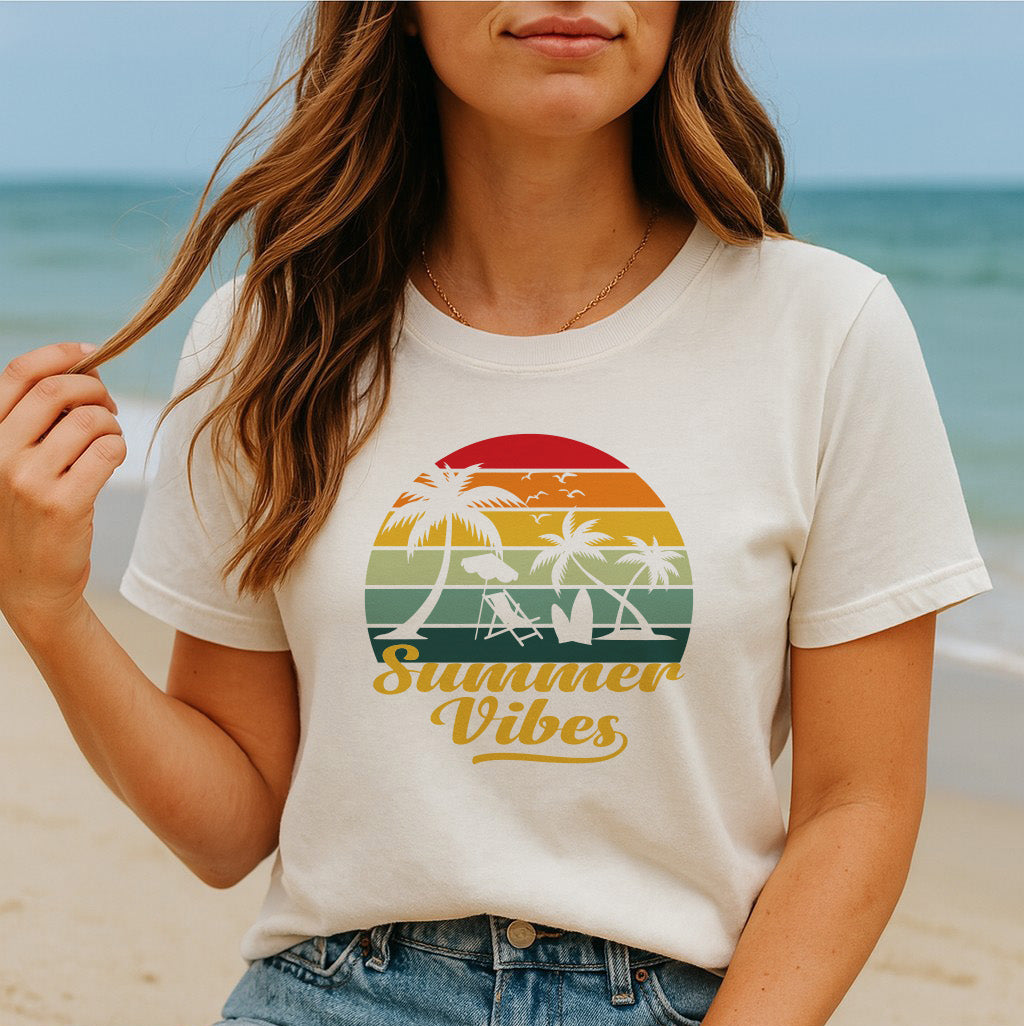 Women's Summer Vibes graphic tee with retro sunset and palm trees design, worn seated on the beach.