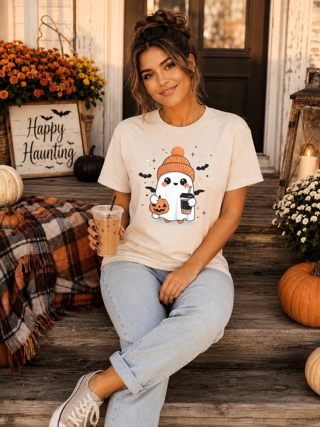 A woman sits on rustic wooden porch steps wearing a cream-colored graphic t-shirt with a cute cartoon ghost wearing an orange beanie and holding a pumpkin. The background is decorated for Halloween with pumpkins, flowers, and a 'Happy Haunting' sign.