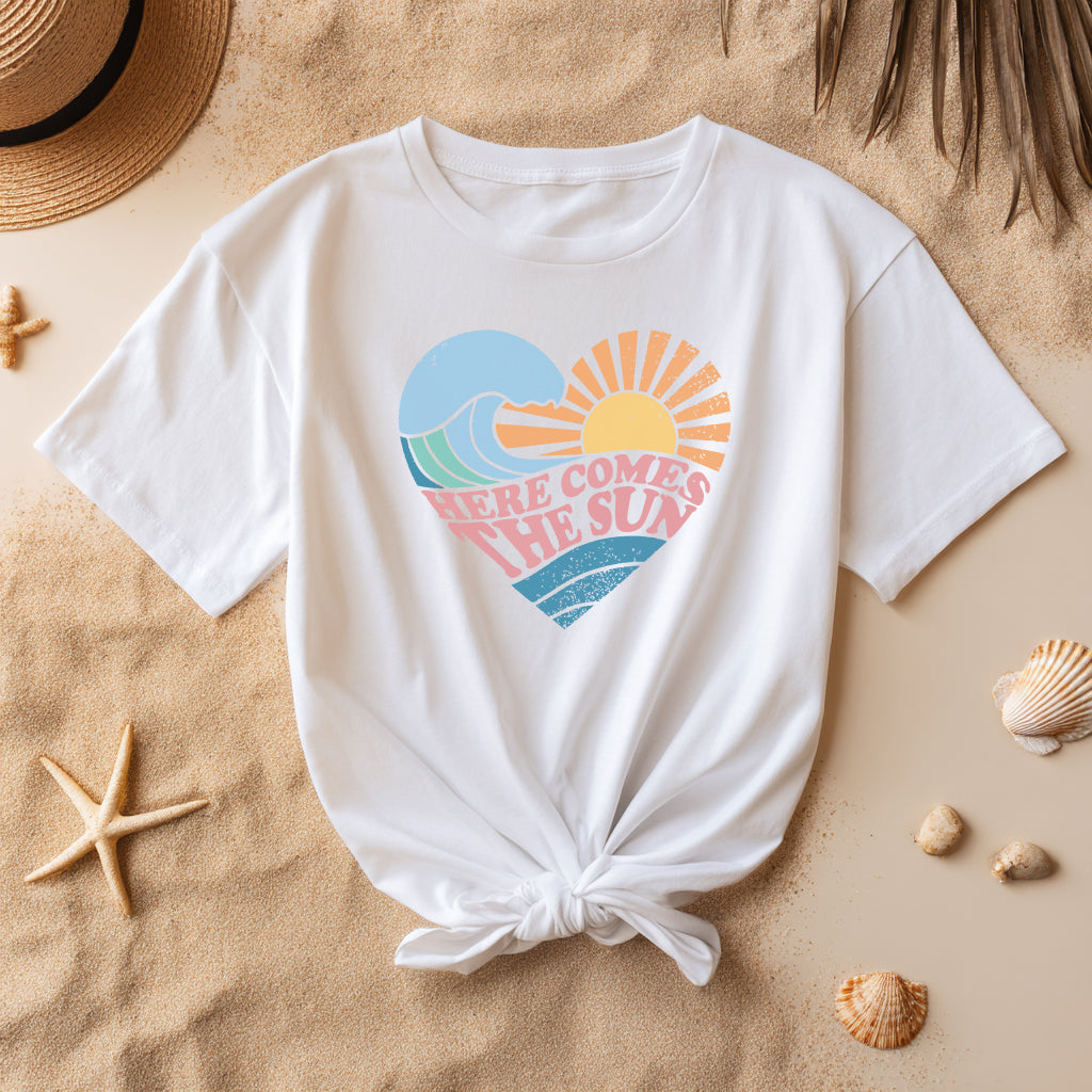 Beach flatlay of white retro graphic tee tied at waist, featuring pastel heart design with sun and waves and text ‘Here Comes The Sun.
