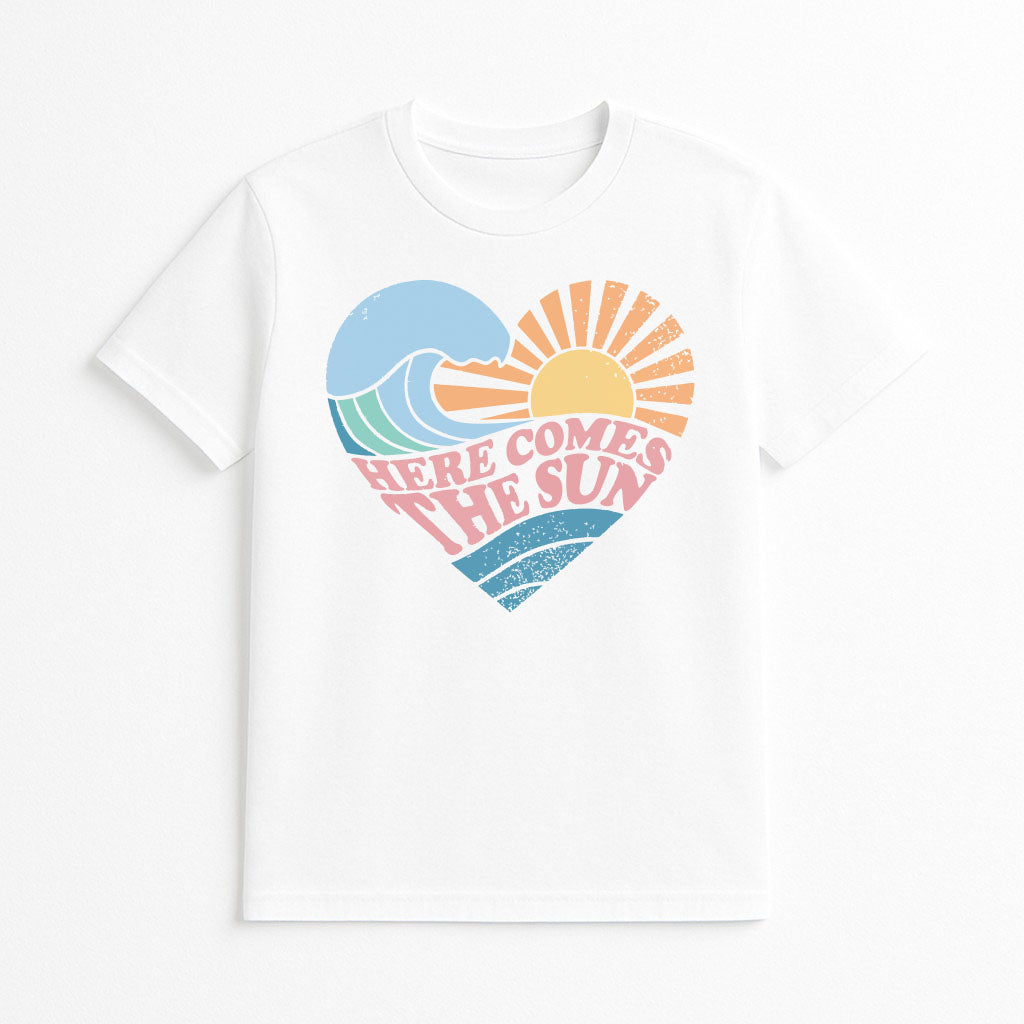Studio flatlay of white women's graphic tee with retro heart design reading ‘Here Comes The Sun’ in pastel waves and sunshine illustration.
