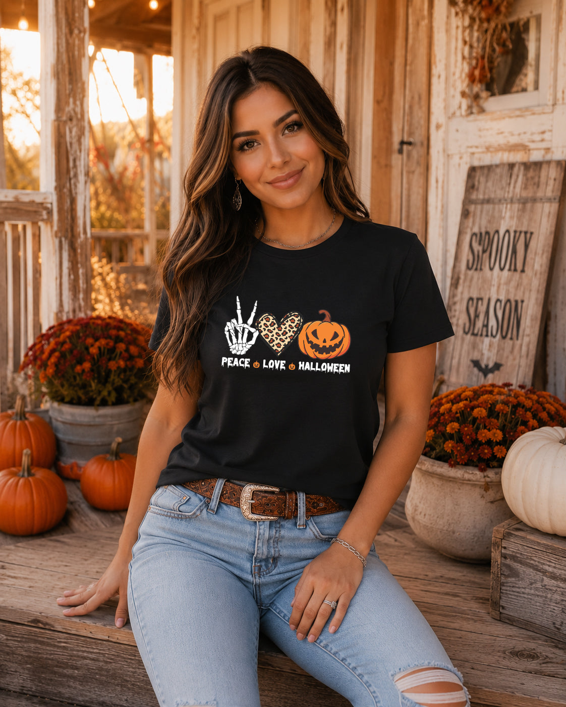 Woman wearing the Peace Love Halloween graphic tee featuring a skeleton peace sign, leopard print heart, and jack-o'-lantern pumpkin while relaxing on a rustic fall porch with pumpkins and autumn décor.