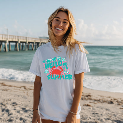 Woman wearing the Hello Summer crab graphic tee on a beach featuring a colorful coastal design with crab, starfish, seashells, sunglasses, and tropical summer elements.