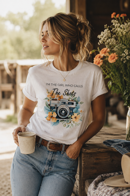 Woman wearing the I'm The Girl Who Calls The Shots photographer white graphic tee featuring a vintage camera with floral artwork, styled with western-inspired denim in a rustic setting.